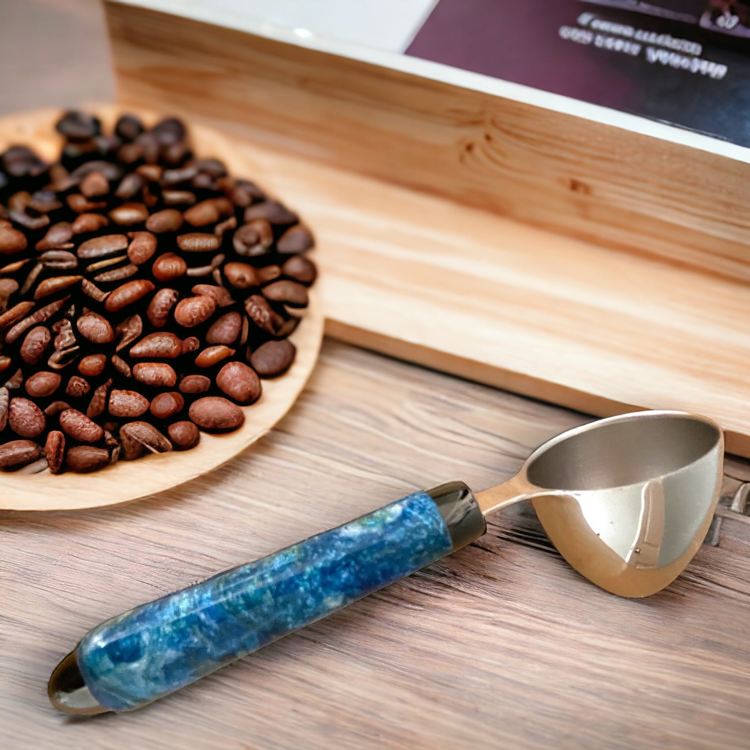 Picture of a blue, green, and white hand turned coffee scoop sitting on a counter with a bowl of coffee beans made by Paul’s hand turned creations. 