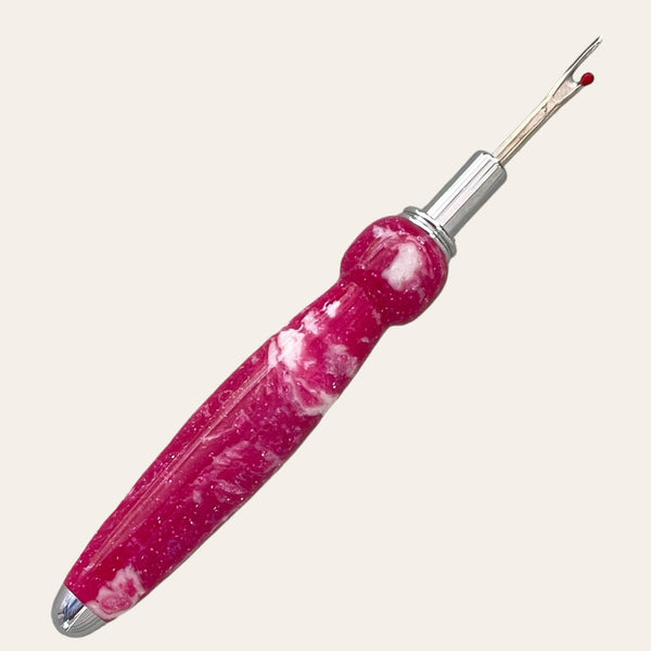 The Magenta Resin Single Blade Seam Ripper by Paul's Hand Turned Creations is a handmade tool featuring a bright pink, sparkly handle and a sharp metal tip for cutting stitches in fabric. This essential sewing tool has an ergonomic rounded end and includes a small red ball on one prong for safety.