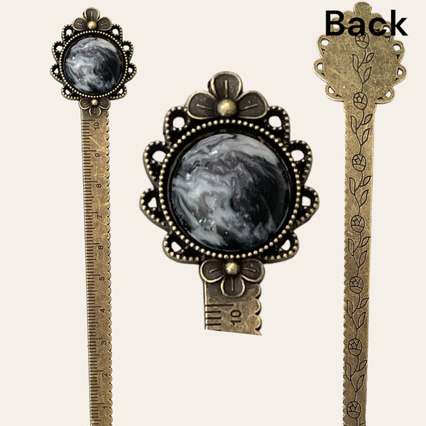 The "Black & White Resin Hand Turned Cabochon Bookmark" from Paul's Hand Turned Creations features ornate antique brass with intricate floral designs. The top of the bookmark boasts a black and white marble-like resin cabochon set in lace-like filigree. Similar floral detailing is displayed on the back. Additionally, a ruler in both inches and centimeters is incorporated into the shaft, making it a perfect blend of beauty and utility.