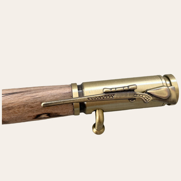 Close-up image of the 30 Caliber Rifle Bolt Action Pen with Antique Brass - Spalted Maple Wood by Paul's Hand Turned Creations. The clip is designed to resemble a .30 caliber rifle with intricate engravings, making it perfect for hunting enthusiasts. The wooden barrel of this handmade pen showcases the natural grain patterns of spalted maple wood. This pen's design is both intricate and rustic, seamlessly blending antique brass metal elements with natural wood.