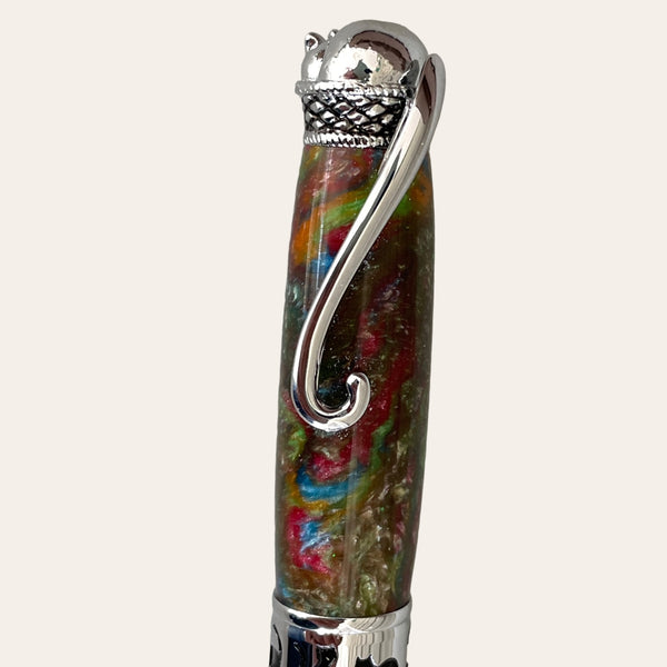 Close-up of the Hand Turned Cat Pen on Chrome Trim- Calico by Paul's Hand Turned Creations, showcasing a colorful, marbled barrel with red, blue, green, and yellow hues. This hand-turned cat pen features intricate silver accents, including a decorative cap and a sturdy curved clip. The background is plain and light-colored.