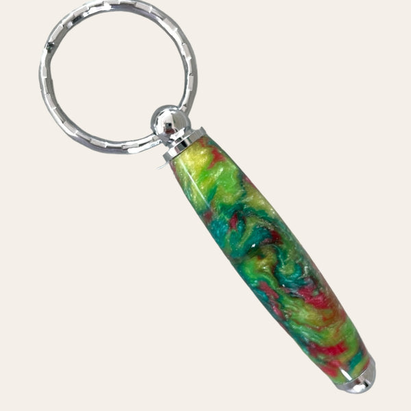 The Small Resin Keychain - Mysterious by Paul's Hand Turned Creations is a handcrafted piece featuring a metal ring and a colorful resin handle. The handle showcases an abstract swirl pattern in vibrant shades of green, yellow, red, and blue. The polished metal components offer a stunning contrast to the vivid colors of the resin, making it a unique and eye-catching accessory.