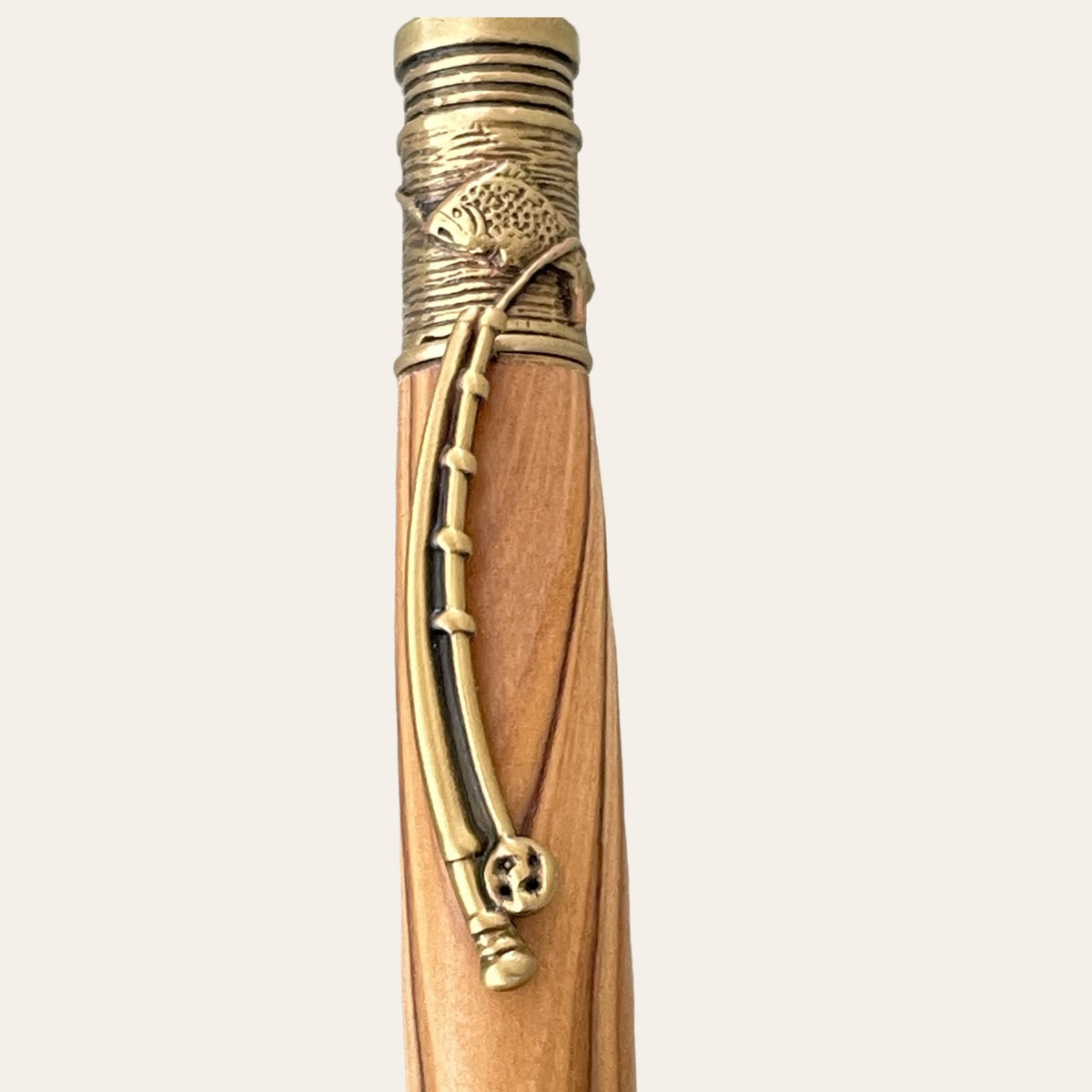 A close-up view of the top segment of the Exotic Bethlehem Olive Wood Fly Fishing Pen With Antique Brass Trim by Paul's Hand Turned Creations. The pen features an intricate metal trim with an ornate design, including a fish and a curved line resembling a fishing pole engraved on the antique brass part. The background is plain white.