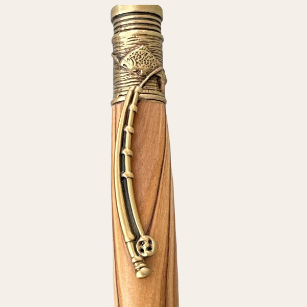 A close-up view of the top segment of the Exotic Bethlehem Olive Wood Fly Fishing Pen With Antique Brass Trim by Paul's Hand Turned Creations. The pen features an intricate metal trim with an ornate design, including a fish and a curved line resembling a fishing pole engraved on the antique brass part. The background is plain white.