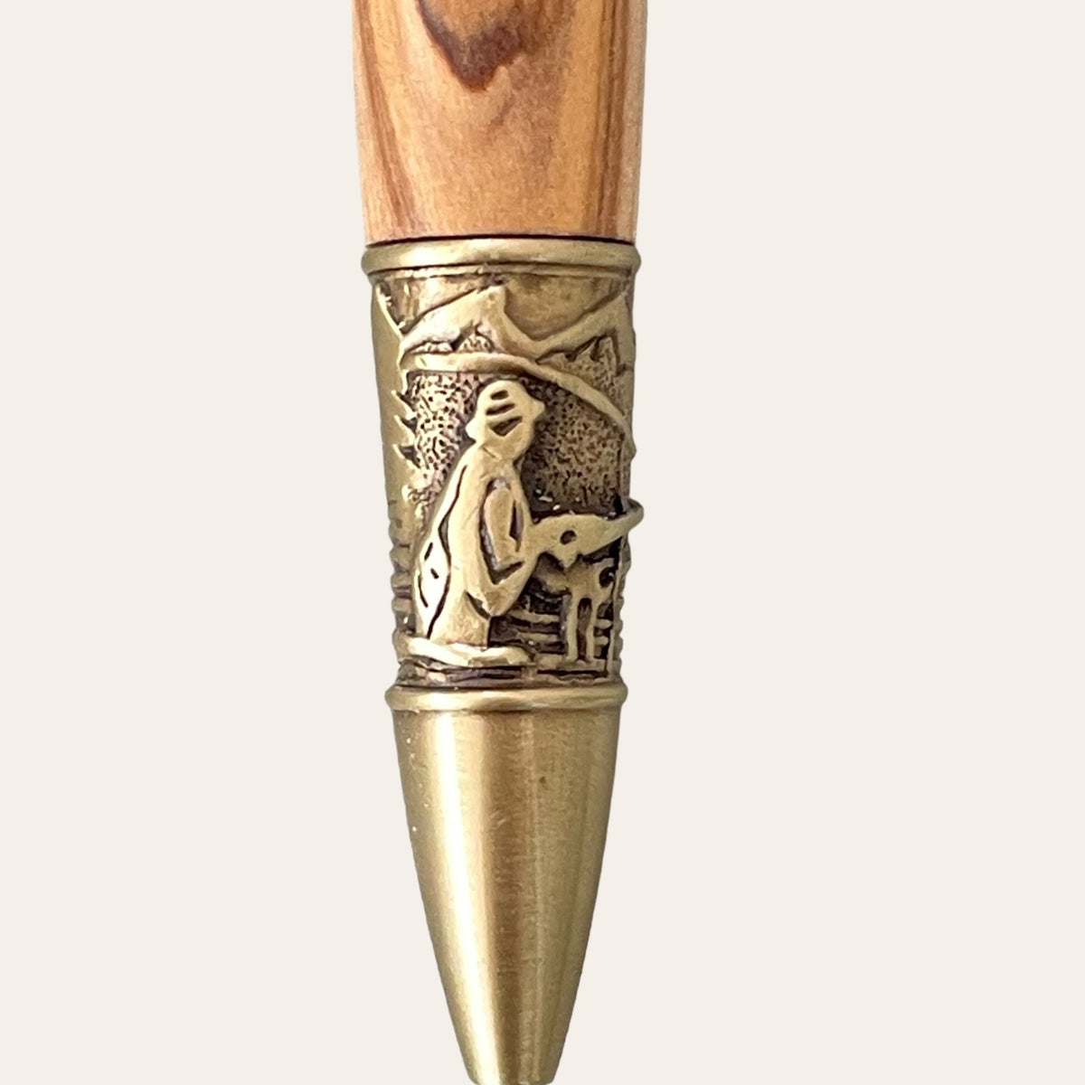 Close-up of the tip of Paul's Hand Turned Creations' Exotic Bethlehem Olive Wood Fly Fishing Pen with Antique Brass Trim, showcasing a detailed bronze band. This band features an intricate design of a fisherman holding a rod, set against scenic mountains and a river in the background. The natural grain pattern of the handcrafted olive wood above the band is complemented by the antique brass refillable pen kit.