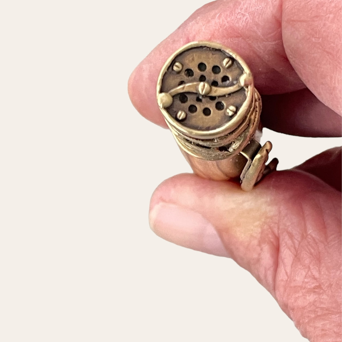 A close-up of a person's hand holding a small, cylindrical, vintage-looking object adorned with antique brass trim from Paul's Hand Turned Creations. Called the Exotic Bethlehem Olive Wood Fly Fishing Pen, it features a perforated pattern on the top for sound or air passage and some curved decorative elements. The background is plain and off-white.