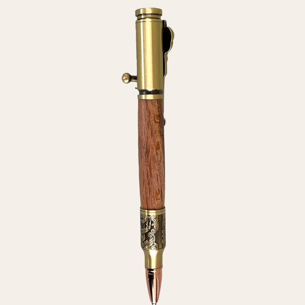 A vintage-style pen named "Hand Turned Deer Hunter Bolt Action with Antique Brass - Laurel Oak Wood" by Paul's Hand Turned Creations, featuring a wooden body and antique brass finish. It includes decorative carvings near the tip and a matching clip at the top. This handmade pen boasts intricate detailing, giving it an elegant, classic look against a plain and light-colored background.