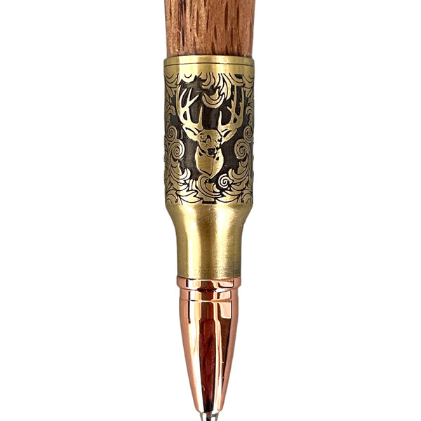 A close-up of the Hand Turned Deer Hunter Bolt Action with Antique Brass - Laurel Oak Wood pen by Paul's Hand Turned Creations showcases its detailed, ornate design. This handmade pen features an intricately engraved brass section depicting a deer with antlers surrounded by swirling patterns. The writing tip is metallic with a copper finish, and the upper part is crafted from laurel oak wood.