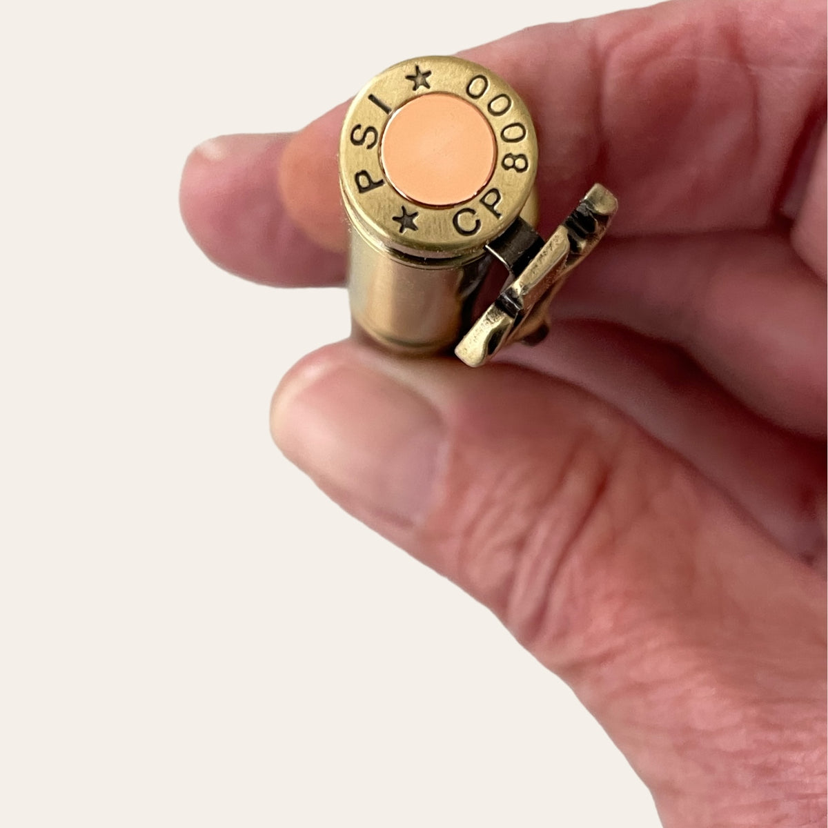 A close-up of a hand holding a brass cartridge case with an antique brass finish and a primer marked "PSI * 0008 * CP". The cartridge is snugly gripped between the thumb and index finger against a neutral background, reminiscent of the craftsmanship in creating Paul's Hand Turned Creations' Hand Turned Deer Hunter Bolt Action with Antique Brass - Laurel Oak Wood pen.