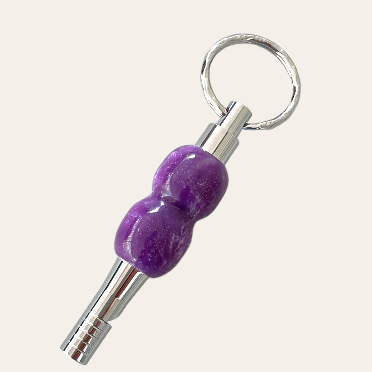 The Lilac Resin Key Chain with Safety Whistle by Paul's Hand Turned Creations features a metal keychain with a circular ring holder. It boasts a metallic base, an ergonomically shaped purple grip in the center, and a ridged metallic tip. The purple grip includes two rounded sections, giving it a curvy appearance. This unique accessory doubles as a safety whistle for added convenience.