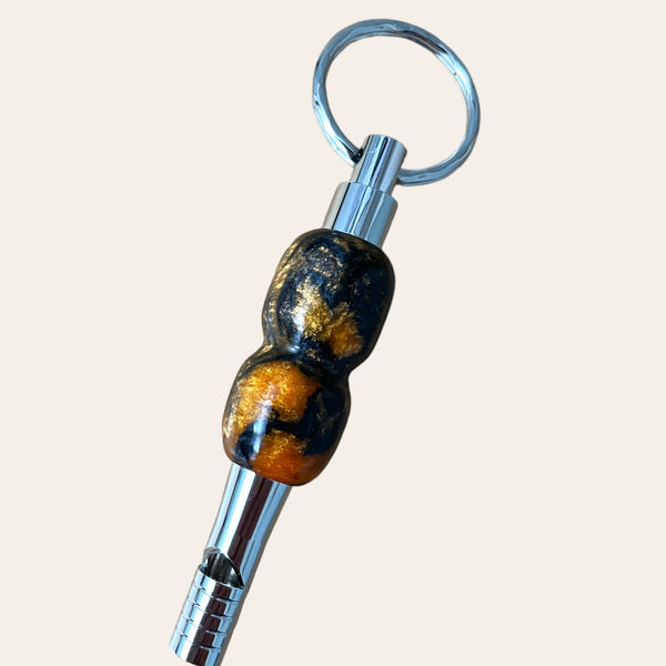 A Black and Orange Resin Key Chain with Safety Whistle by Paul's Hand Turned Creations features a polished keychain with a sleek silver whistle and decorative bead. The bead boasts a glossy finish, showcasing a captivating blend of black and golden hues. The whistle is elegantly attached to the metallic ring, making it both a practical accessory and a unique gift idea.