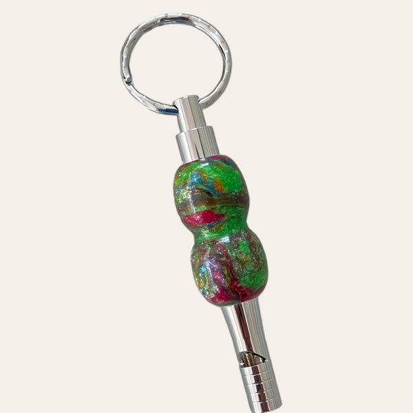 Check out the Multi-Colored Resin Key Chain with Safety Whistle by Paul's Hand Turned Creations. This unique accessory combines a marbled green and red handcrafted centerpiece, resembling a bead, attached to a silver safety whistle and finished with a keyring at the top. The background is off-white.