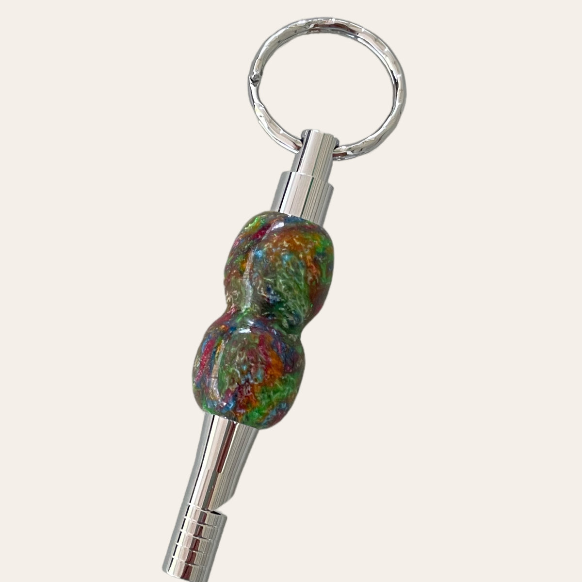 Paul's Hand Turned Creations presents the Multi-Colored Resin Key Chain with Safety Whistle, featuring a sleek silver metallic loop at the top and an abstract-shaped, colorful resin element in the center. The vibrant blend of green, red, blue, and gold hues creates an eye-catching look that combines modern design with lively colors.