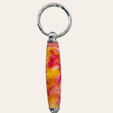 A close-up photograph showcases the Paul's Hand Turned Creations' Small Resin Keychain - Lotus Flower. This handcrafted piece features a cylindrical handle with a marbled design in vibrant shades of pink, yellow, and white, reminiscent of delicate lotus flower petals. The keychain is elegantly attached to a silver metal ring, set against a plain light beige background.