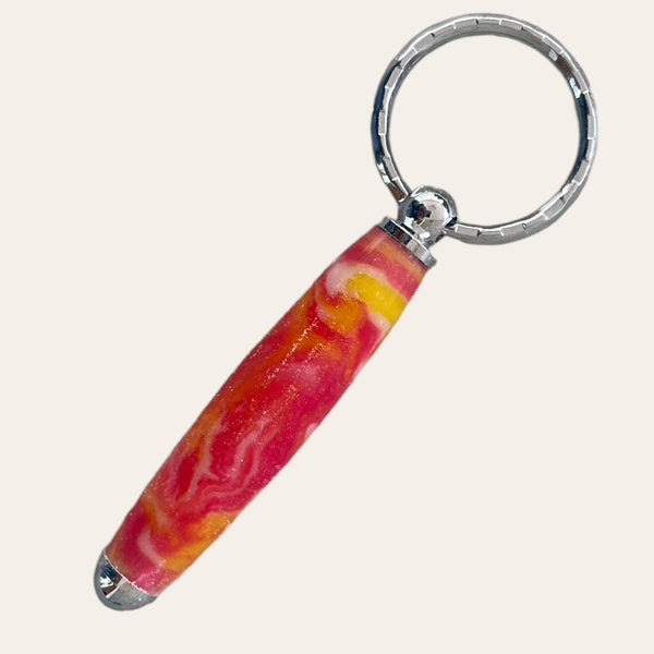 A close-up of the Small Resin Keychain - Lotus Flower by Paul's Hand Turned Creations, showcasing its cylindrical, marbled pink and yellow handle with metallic silver ends. The handle is attached to a round, metallic key ring. The plain background emphasizes the colorful detail of this handcrafted accessory.

