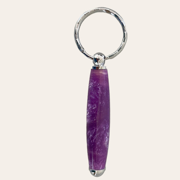 A silver, handcrafted keychain from Paul's Hand Turned Creations, featuring a lilac, cylindrical attachment known as the Small Resin Keychain - Lilac, is displayed against a plain, light background. The lilac attachment boasts a smooth, slightly glossy finish with delicate marbling patterns. This unique keychain adds a touch of elegance to any set of keys.