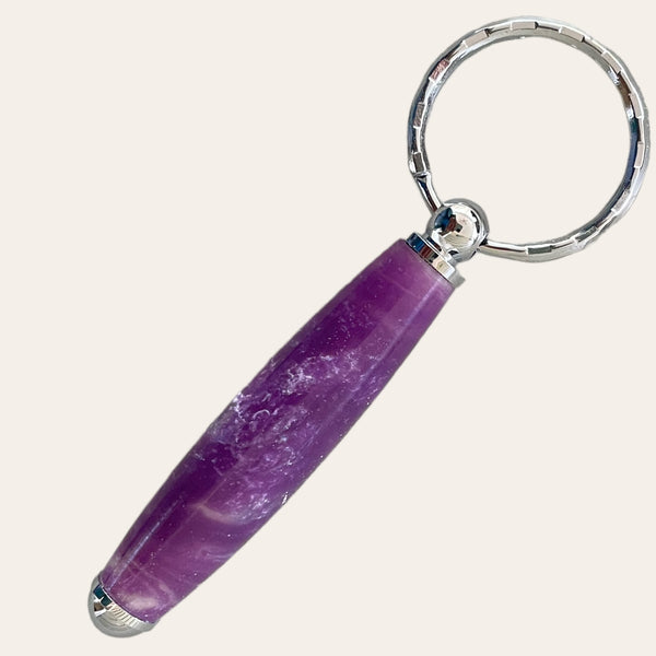 A handcrafted keychain from Paul's Hand Turned Creations, named the Small Resin Keychain - Lilac, features a large, cylindrical purple stone adorned with lighter marbled patterns and a polished finish. The shiny and reflective metal parts attaching the stone to the silver ring make this unique keychain stand out against the plain white background.