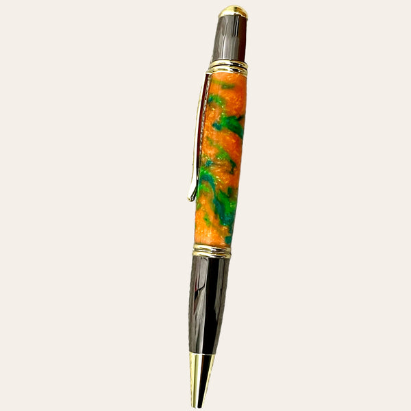 A close-up of a Hand Turned Gatsby Refillable Pen - Harmony from Paul's Hand Turned Creations, showcasing an orange and green marbled barrel. The pen's unique design includes shiny metallic accents in gold and chrome on the tip, clip, and cap ends. This vibrant writing instrument is vertically aligned against a neutral beige background.