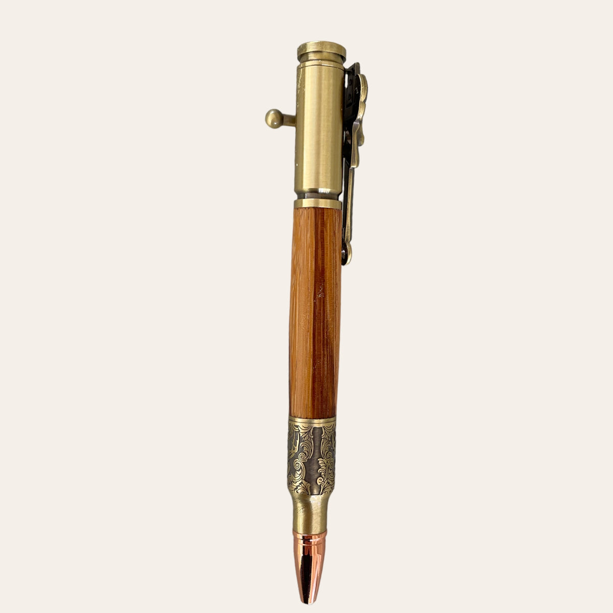 The Hand Turned Deer Hunter Bolt Action with Antique Brass - Zebrawood from Paul's Hand Turned Creations features a metallic gold-colored bolt action mechanism and clip at the top, complemented by a wooden grip made of Zebrawood in the middle. The pen's ornate tip showcases intricate engravings and a copper-colored nib resembling a bullet, all set against a plain cream background.