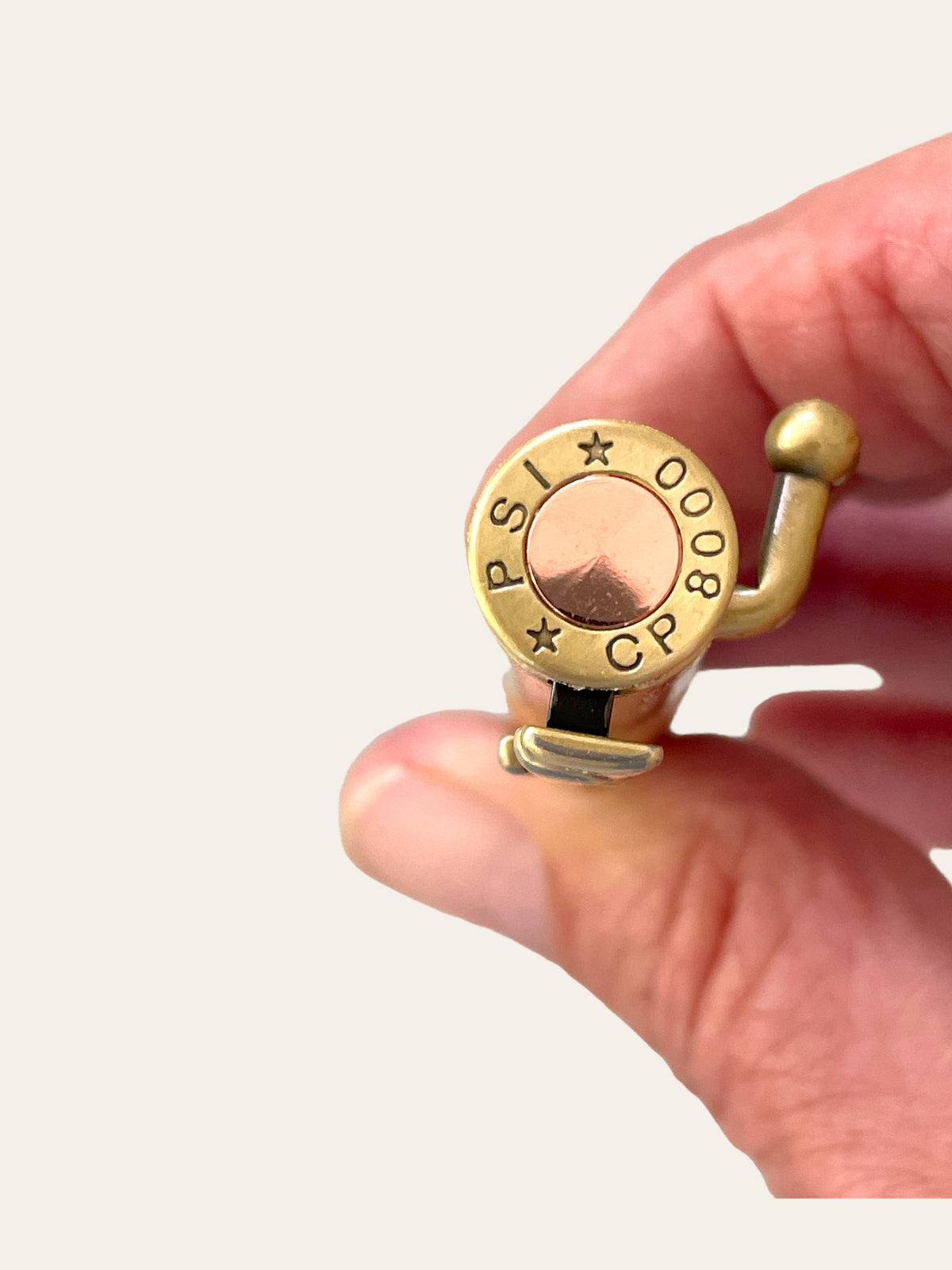 Close-up of a hand holding an antique brass-colored circular object with an engraved top featuring the text "PSI 00008 CP." The object, resembling a 30 Caliber Rifle Bolt Action Pen, has a small handle-like extension on one side. The background is plain and white. This unique creation is part of Paul's Hand Turned Creations collection, specifically highlighting the spalted tamarind material.