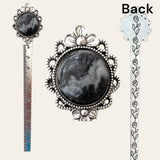 The Black and White Hand Turned Cabochon Resin Bookmark by Paul's Hand Turned Creations features a silver finish with a black and white gemstone at the top, encased in intricate floral metalwork. The back displays an engraved pattern, and this antique-style silver bookmark also functions as a ruler along its length.