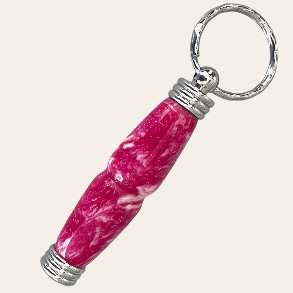 The Magenta Hand Turned Resin Secret Compartment Key Chain from Paul's Hand Turned Creations features a metallic ring with a screw cap next to a pink marbled cylindrical object with threaded ends, indicating that the components can be assembled together. The background is a plain off-white color.