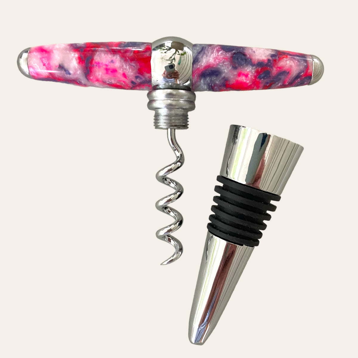 A vibrant, handmade creation from Paul's Hand Turned Creations, the Bubblegum Resin Hand Turned T-Handle Winetopper Corkscrew, features a bright pink and purple swirled handle. Next to it lies a shiny silver wine topper adorned with black rubber rings. The corkscrew boasts a metallic spiral and a reflective, bulbous center. Both items are set against a plain, light background.