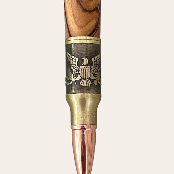 A close-up of the Bald Eagle Bolt Action Hand Turned Pen with Bethlehem Olive Wood by Paul’s Hand Turned Creations showcases a detailed engraving of an eagle with outspread wings, clutching arrows and an olive branch, flanked by a shield and stars. This patriotic writing tool features a wooden texture and antique brass finish, giving it a refined and patriotic appearance.