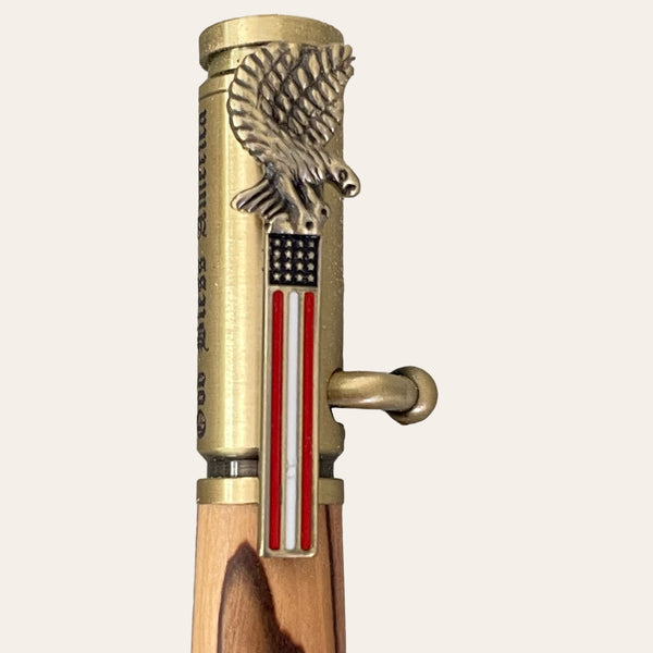 Introducing the Bald Eagle Bolt Action Hand Turned Pen with Bethlehem Olive Wood, a patriotic writing instrument from Paul's Hand Turned Creations. This pen boasts an intricate design with a brass clip adorned by a bald eagle perched atop an American flag. The body showcases a wooden appearance, complemented by antique brass finish accents for an added touch of sophistication.