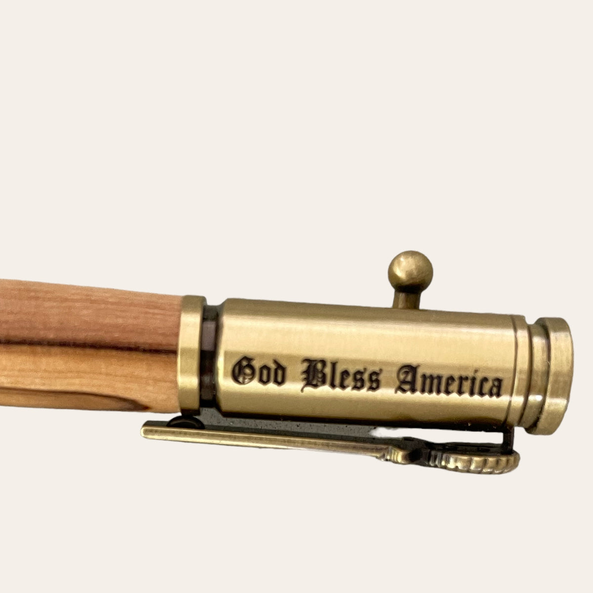 A close-up image showcases the metallic clip of a Bald Eagle Bolt Action Hand Turned Pen with Bethlehem Olive Wood. This intricately designed clip from Paul's Hand Turned Creations, featuring an antique brass finish, bears the engraving "God Bless America" and mirrors the bolt action mechanism of a rifle, making it an exceptional patriotic writing tool.