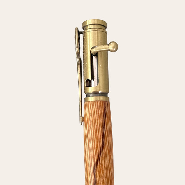 A close-up of the 30 Caliber Rifle Bolt Action Pen with Antique Brass - Zebrawood from Paul's Hand Turned Creations. The pen features a brass upper section boasting a unique bolt-action mechanism reminiscent of a rifle, along with a built-in pocket clip, and a beautifully grained zebrawood lower section. It is positioned vertically against a plain, off-white background.