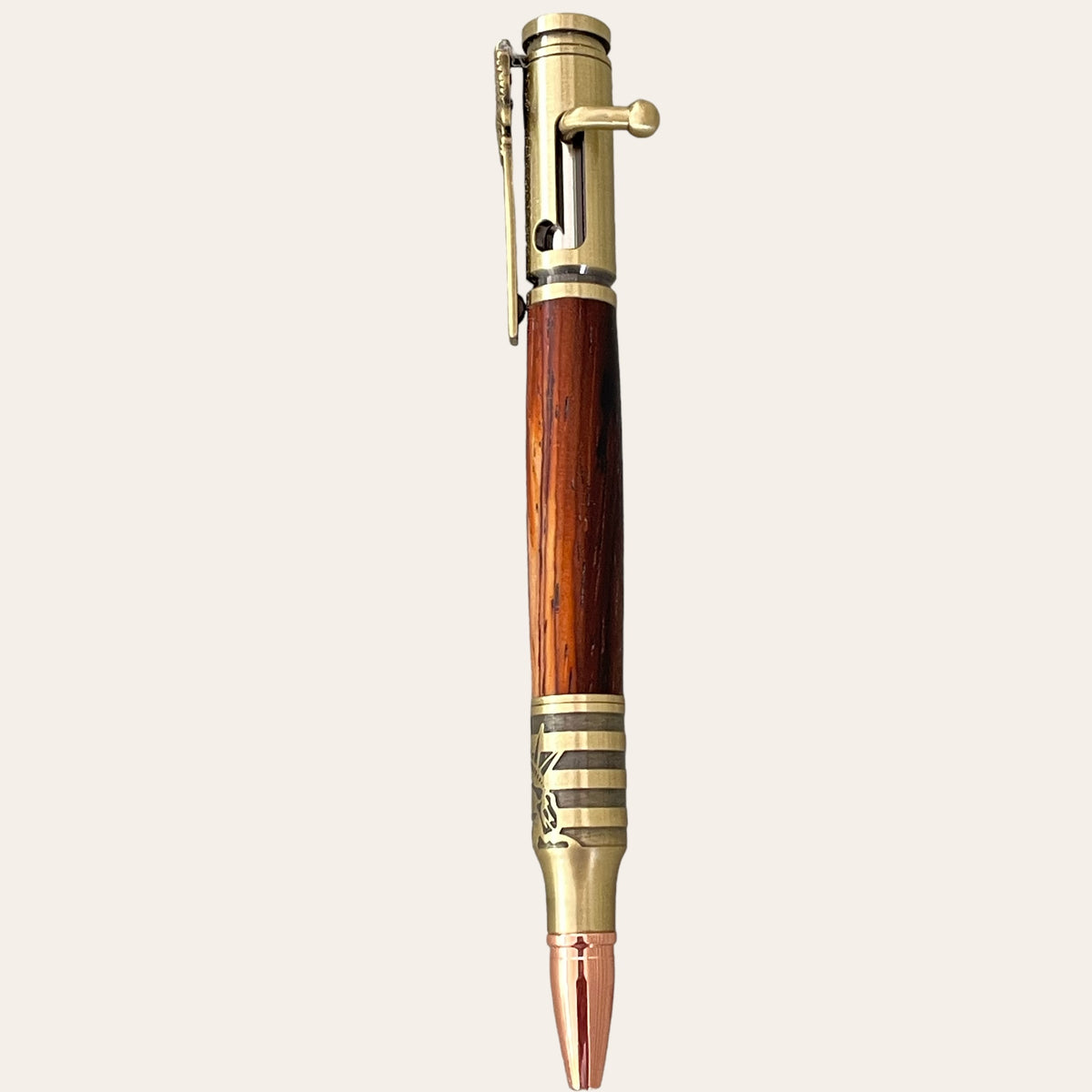 The Statue Of Liberty Bolt Action Hand Turned Pen by Paul's Hand Turned Creations features a Bocote wood barrel and antique brass accents. The top part includes a bolt-action mechanism, and the tip resembles a bullet. This patriotic writing tool has a metal clip, adding to its rustic and vintage aesthetic against a plain beige background.