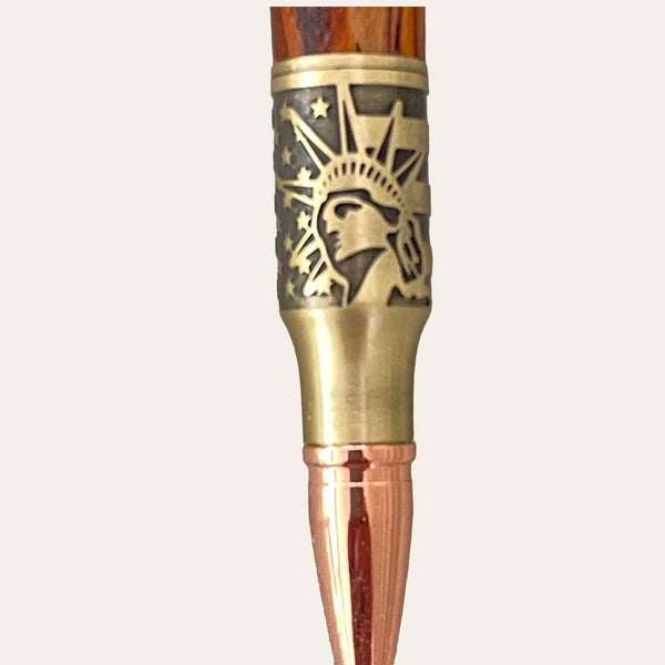 A detailed close-up of the Statue Of Liberty Bolt Action Hand Turned Pen - Bocote Wood from Paul's Hand Turned Creations showcases an intricate metal design near the top, featuring the Statue of Liberty against a backdrop of stars. The pen features a wooden body with an antique brass finish and a polished metallic tip.