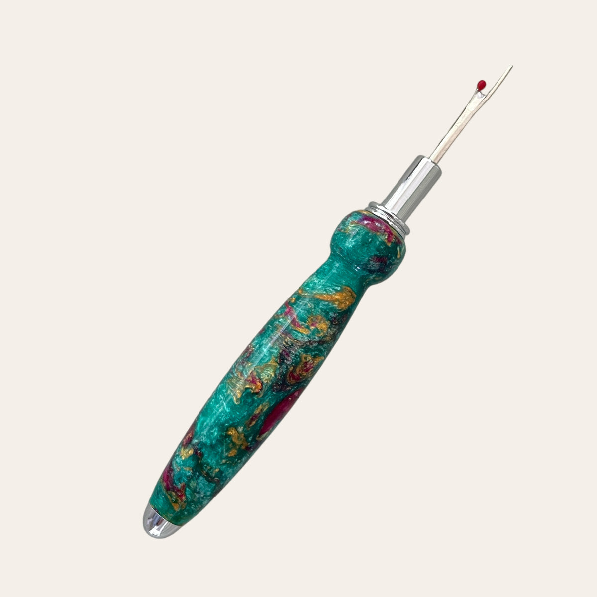 The Royal Jewels Resin Single Blade Seam Ripper from Paul's Hand Turned Creations is an essential sewing tool with a vibrant, colorful marbled handle featuring shades of green, red, and gold. It boasts a stainless steel blade and a metal tip equipped with a small red ball for cutting threads. The background is light beige.