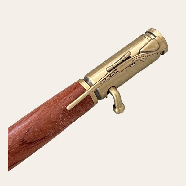 A close-up image of the Bubinga Wood 30 Caliber Rifle Bolt Action Pen with Antique Brass Trim by Paul's Hand Turned Creations, showcasing its elegant wooden body and intricate brass details designed to resemble a vintage rifle, complete with a trigger and barrel. This handmade pen uses a Parker Style Refill, set against a plain beige background.