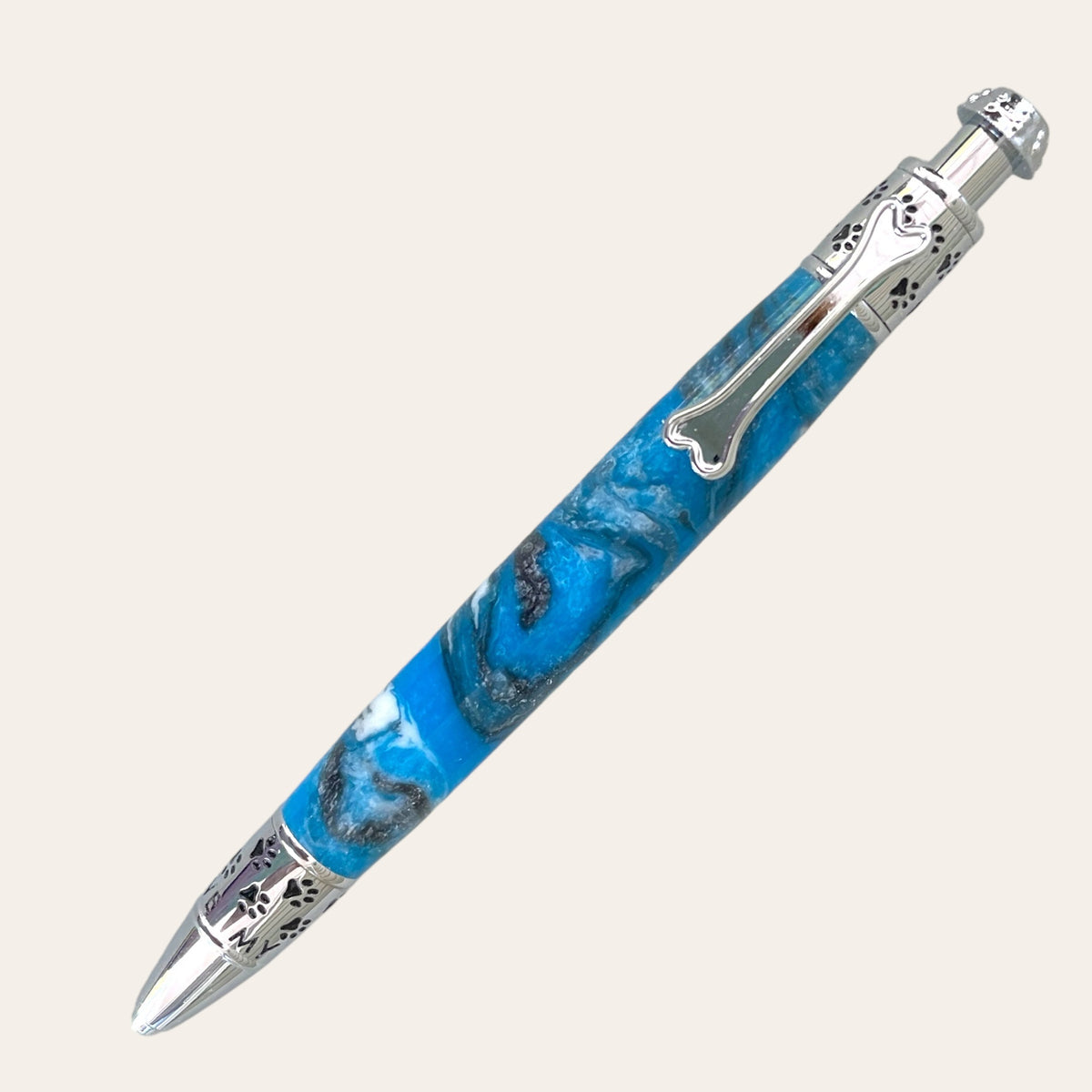 The Resin Dog Click Pen With Chrome Trim - Bluebird, crafted by Paul's Hand Turned Creations, is a stunning blue pen adorned with intricate silver detailing. It features a decorative bone-shaped clip, and its barrel boasts a marbled pattern in shades of blue and white, accented with chrome at both ends. This handcrafted masterpiece is perfect for any dog lover.