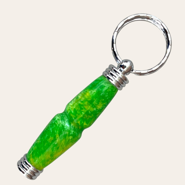 A close-up view of Paul's Hand Turned Creations' Hand Turned Resin Secret Compartment Key Chain - Lemon Lime, showcasing a bright, cylindrical charm. The charm is crafted from durable resin in a primary green color with yellow marbling and features silver metal accents at both ends. The keychain ring itself is metallic and textured. The background is plain and off-white.