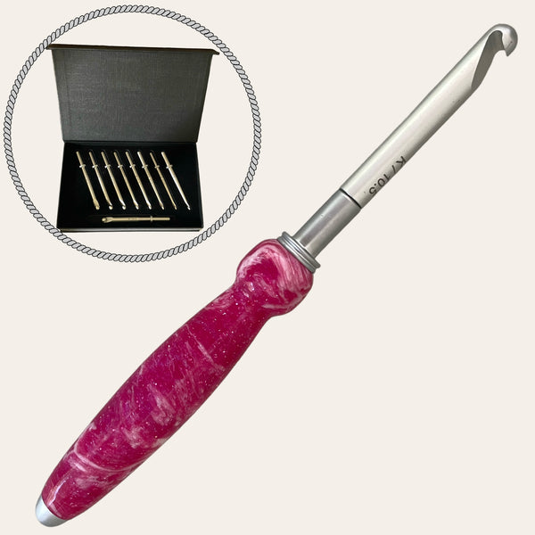 A large pink and white ergonomic crochet hook with a metal tip is shown in front of an inset circle displaying a black box containing Paul's Hand Turned Creations' Hand Turned Resin Crochet Handle with Nine Interchangeable Hooks - Magenta.