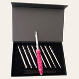 A sleek open case from Paul's Hand Turned Creations contains nine crochet hooks with metal tips, neatly organized inside. One hook, featuring a distinctive pink and white marbled handle, is prominently displayed in the center among the other plain-handled hooks of this elegant magenta crochet set, known as the Hand Turned Resin Crochet Handle with Nine Interchangeable Hooks - Magenta.