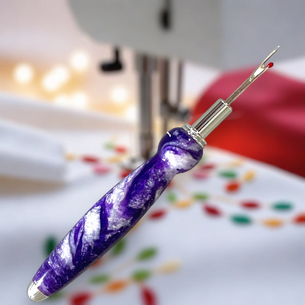 seam ripper made by Paul's Hand Turned Creations with purple and white resin swirls and a stainless steel blade. Background is a muted sewing machine with colorful fabric. 